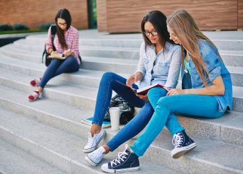 Modern teen girls reading book while sitting on stairs on background of another student