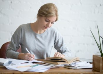 Student female performing a written task in a copybook with a pen, looking at the textbook, education concept photo, horizontal